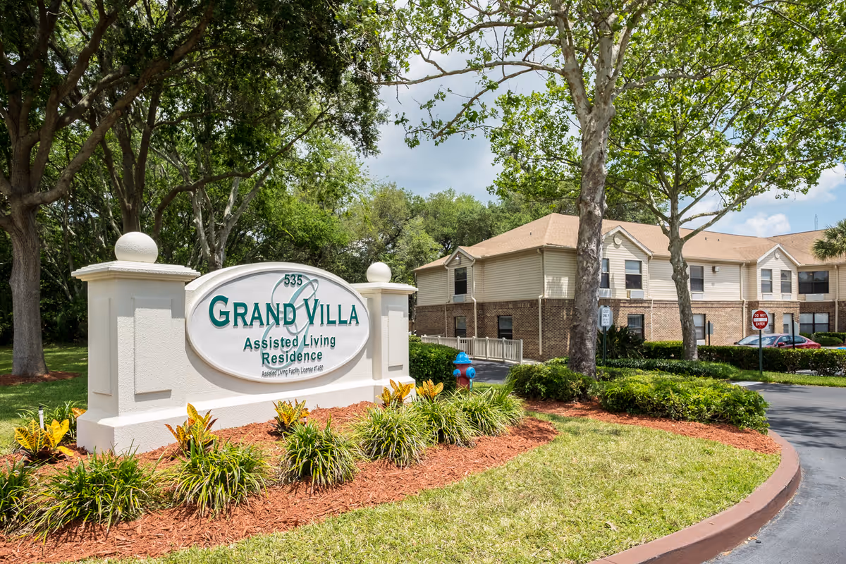 Entrance sign that reads "Grand Villa Assisted Living Residence" surrounded by landscaping with the facility building and trees in the background.