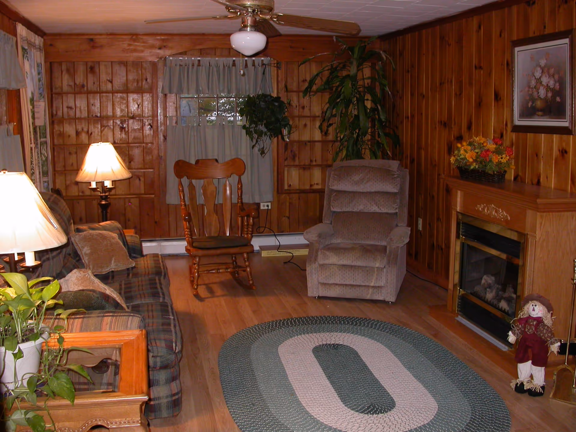 Cozy living room with wood-paneled walls, a plaid sofa with pillows, a wooden rocking chair, a cushioned recliner, a ceiling fan with light, a fireplace with a floral arrangement on the mantel, a small scarecrow decoration, and a braided oval rug on a wooden floor.