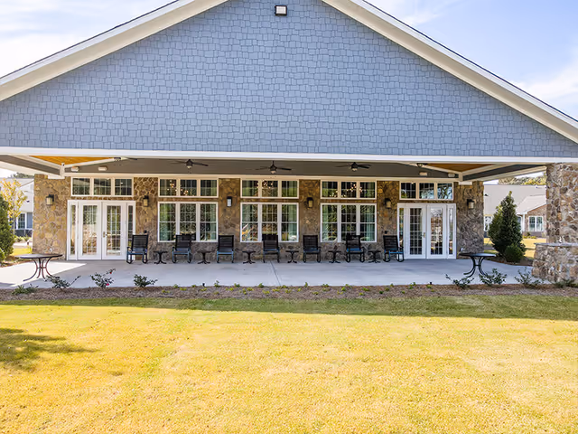 Exterior view of a covered patio area at Manor Lake Hamilton Mill assisted living facility, featuring stone walls, multiple glass doors and windows, ceiling fans, and several black rocking chairs arranged along the patio.