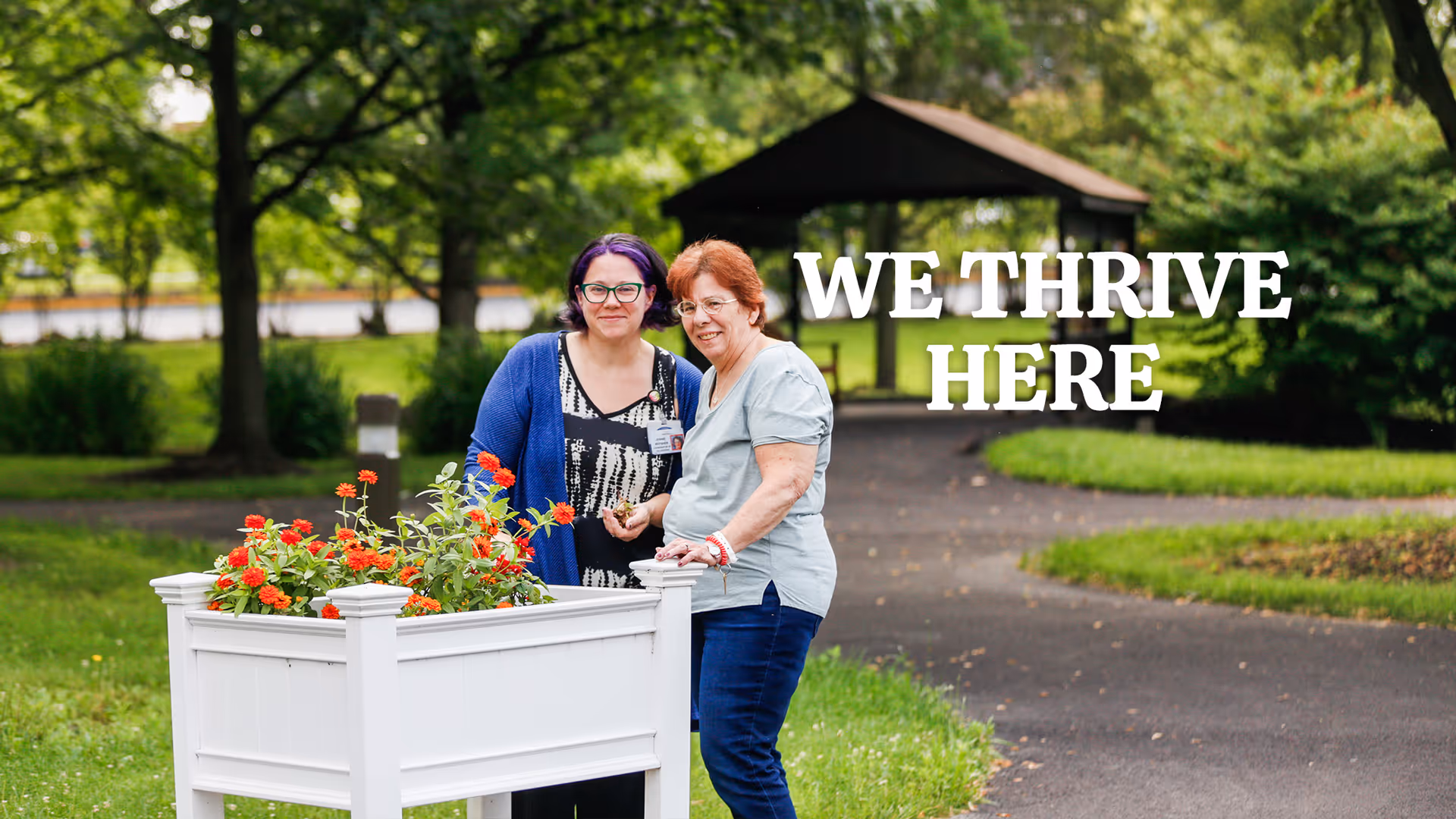Two women standing outdoors near a white planter box filled with red flowers. They are smiling and appear to be enjoying the garden area with trees and a gazebo in the background. The text 'WE THRIVE HERE' is displayed prominently in the image.