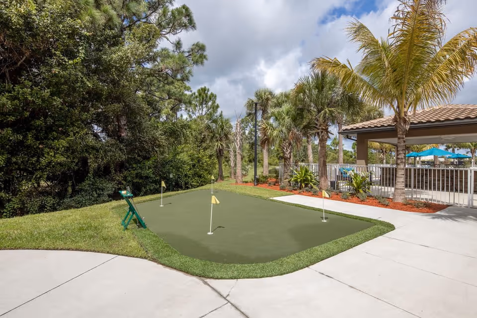 Outdoor putting green with four small flags on holes, surrounded by palm trees and other greenery. There is a covered patio area with blue umbrellas and seating in the background under a partly cloudy sky.
