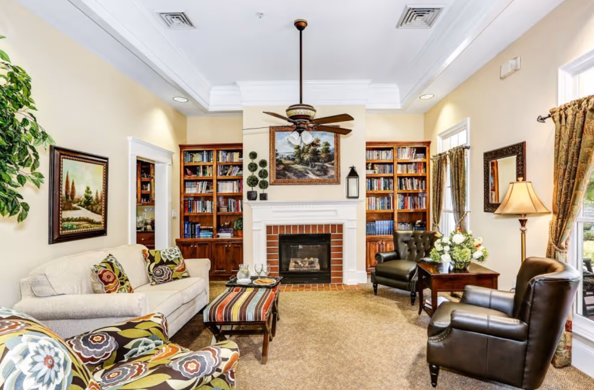 A cozy living room with a beige sofa adorned with patterned cushions, two dark leather armchairs, and a colorful patterned armchair. A striped ottoman serves as a coffee table with a tray holding glasses and a pitcher. The room features a white fireplace with a brick interior, flanked by wooden bookshelves filled with books. Above the fireplace hangs a framed landscape painting. The room is well-lit with natural light coming through windows with patterned curtains, and a ceiling fan with lights hangs from the ceiling.
