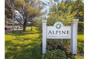 Outdoor view of a grassy area with trees and a white sign that reads 'Alpine Rehabilitation & Nursing Center' under a clear sky.