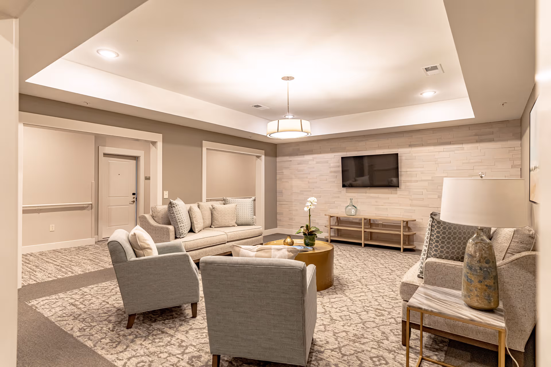 A cozy senior living facility common area with beige and gray tones. The room features a sofa with multiple cushions, two armchairs, a round gold coffee table with a flower vase, a side table with a lamp, and a wall-mounted flat-screen TV on a textured stone accent wall. The carpet has a patterned design, and the ceiling has recessed lighting and a central hanging light fixture.