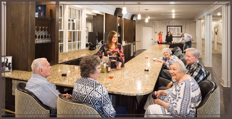 A group of elderly people sitting around a large granite countertop bar in a well-lit room, with a younger woman standing behind the counter serving drinks. In the background, two other people are playing a game at a pool table.