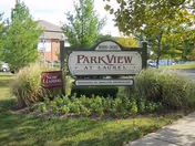 Outdoor view of the entrance sign for Park View at Laurel I, surrounded by greenery and landscaping with trees and bushes, and a smaller sign indicating 'Now Leasing'.