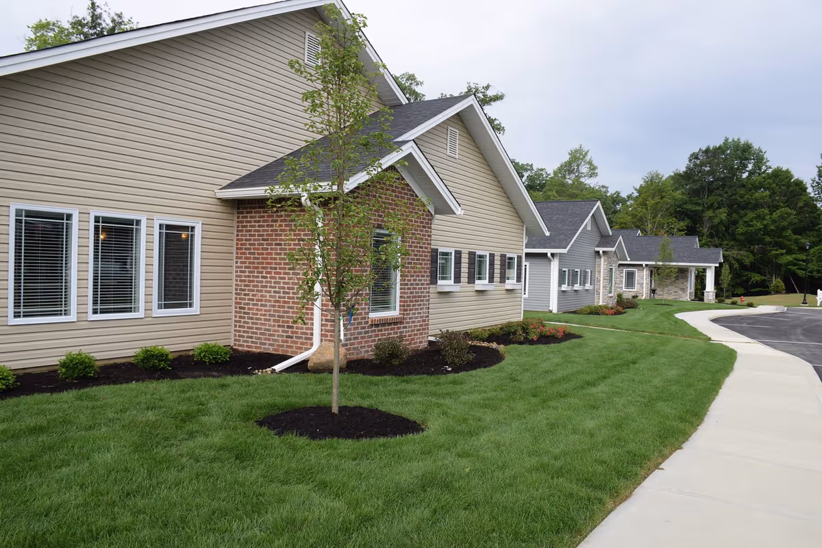 Front exterior of single-story senior living neighborhood buildings with manicured lawn, young trees, and a curved driveway.
