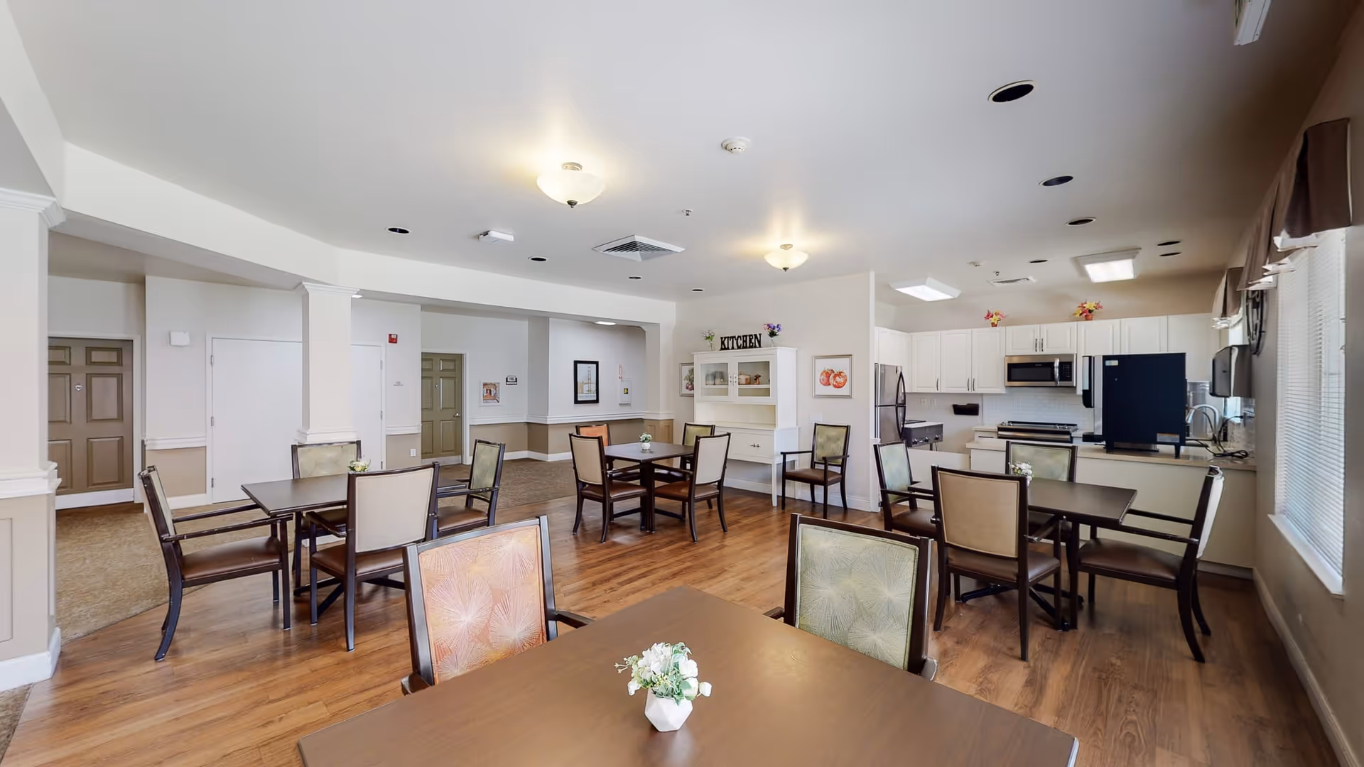 A bright and spacious dining area with several tables and chairs arranged on a wooden floor. The room features a kitchen area with white cabinets, a refrigerator, microwave, and stove. The walls are painted in neutral tones, and there are windows with blinds allowing natural light to enter. Small flower arrangements are placed on the tables.