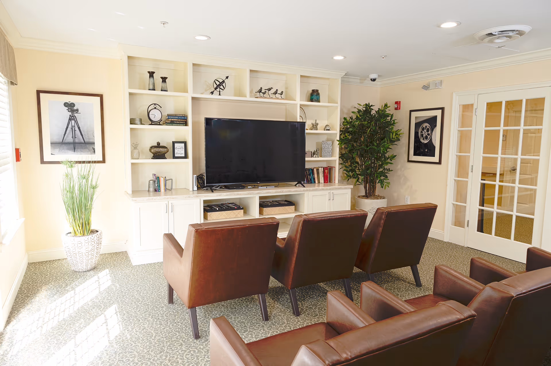 A cozy senior living common area with four brown leather armchairs arranged facing a large flat-screen TV mounted on a white built-in shelving unit. The shelves hold decorative items, books, and baskets. There are two framed black and white photos on the walls, a tall potted plant in the corner, and a window letting in natural light.