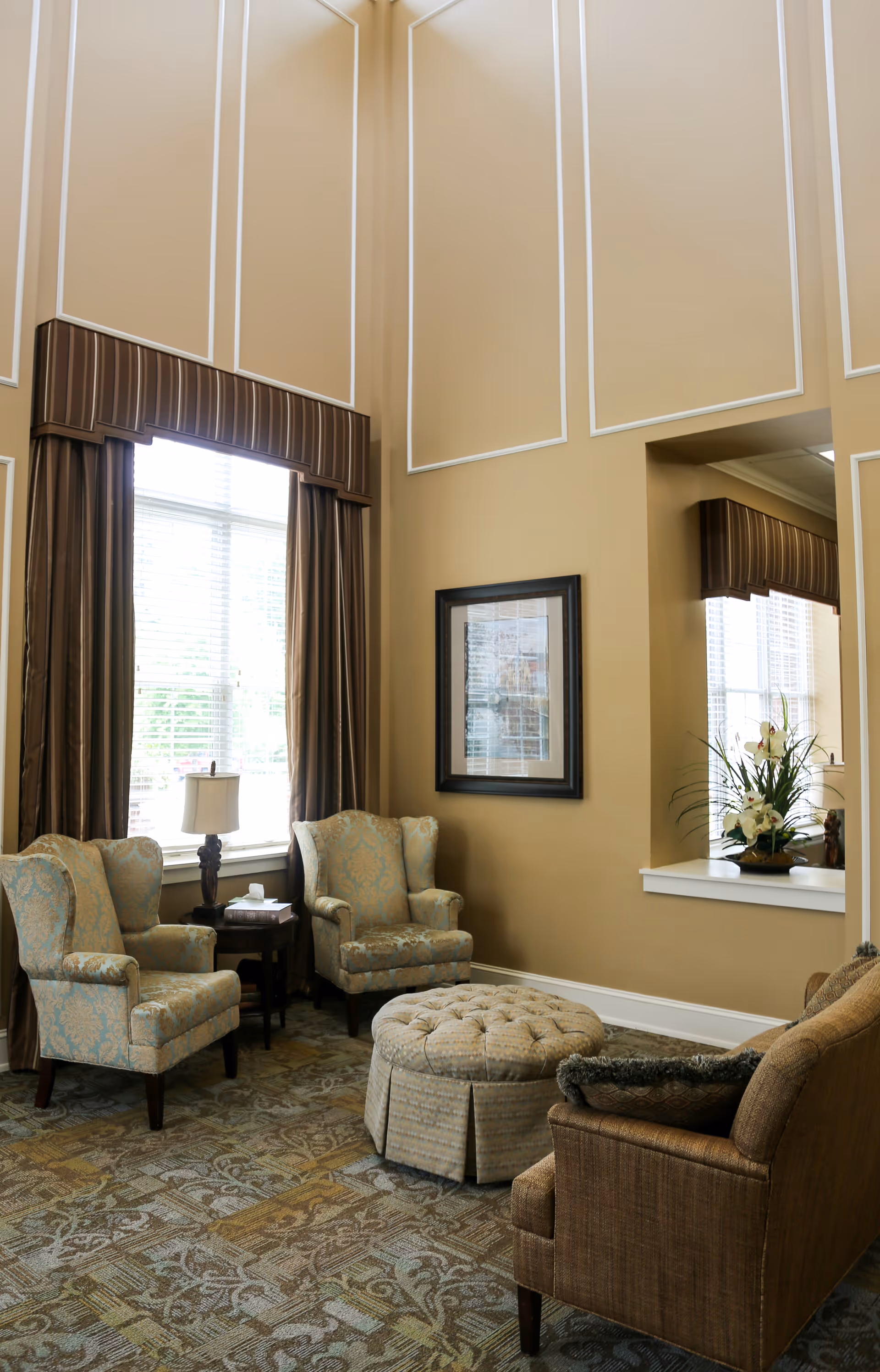 A cozy sitting area with two patterned armchairs, a round tufted ottoman, and a brown upholstered chair. The room features tall beige walls with white trim, large windows with brown curtains, a small side table with a lamp, and a framed picture on the wall. A floral arrangement is placed on a ledge near the window.