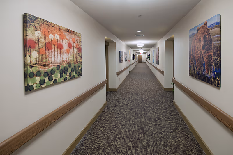 A long, well-lit hallway in a senior living facility with patterned carpet and beige walls. The walls are decorated with colorful paintings, including one of a bear on the right and an abstract floral design on the left. Wooden handrails run along both sides of the hallway.