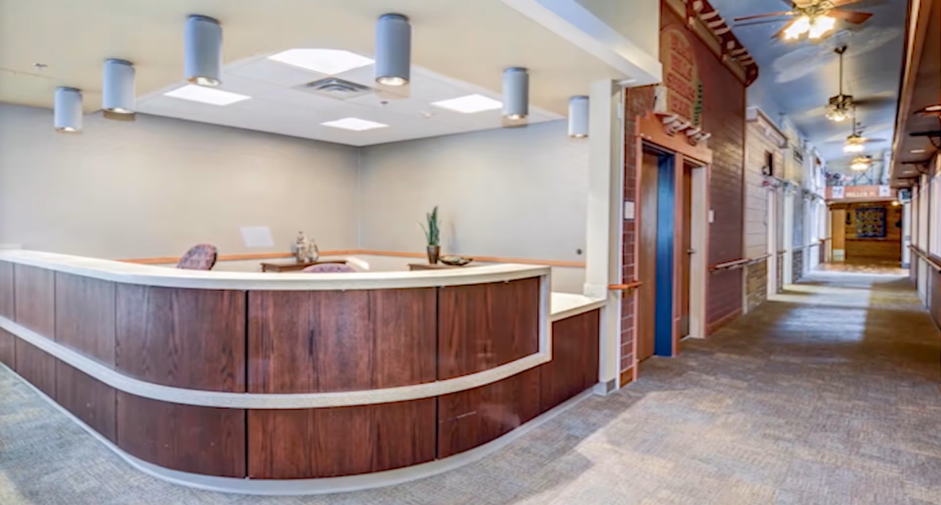 Interior view of a senior living facility reception area with a curved wooden front desk, two chairs behind the desk, a potted plant, and a hallway extending to the right with ceiling fans and handrails along the walls.