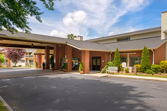 Front exterior view of Brookdale Hendersonville East facility showing a covered entrance with brick pillars, a paved driveway, landscaped greenery including bushes and small trees, and a bench near the entrance under a partly cloudy sky.