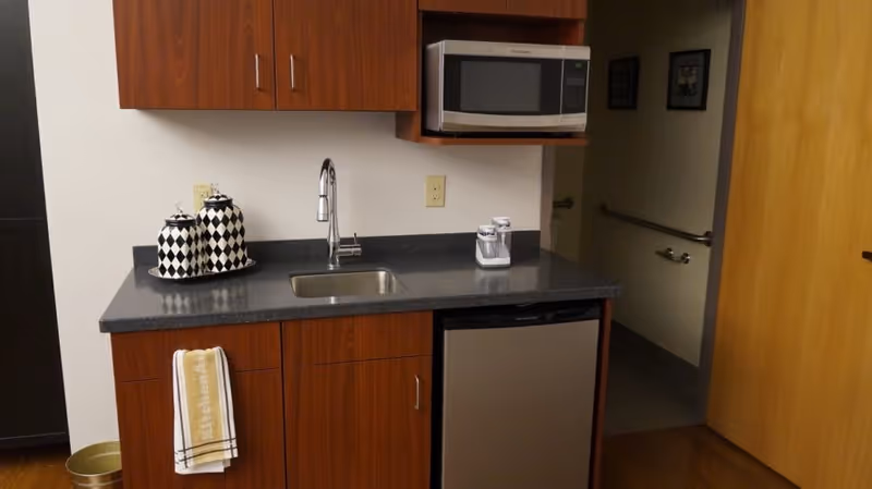 A small kitchenette with wood cabinets, a sink, microwave, mini-fridge and decorative canisters on a dark countertop.