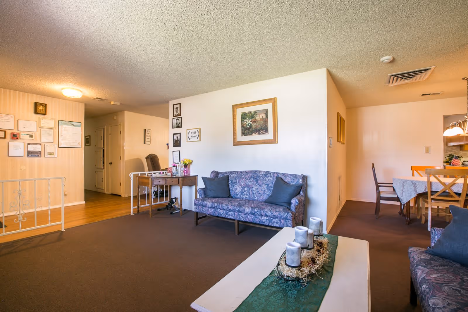 Interior view of a senior living facility lounge area with floral patterned sofas, a white coffee table decorated with candles and a green runner, a wooden side table with flowers and framed pictures on the wall. In the background, there is a dining area with a table covered with a tablecloth and chairs, and a hallway leading to other rooms.