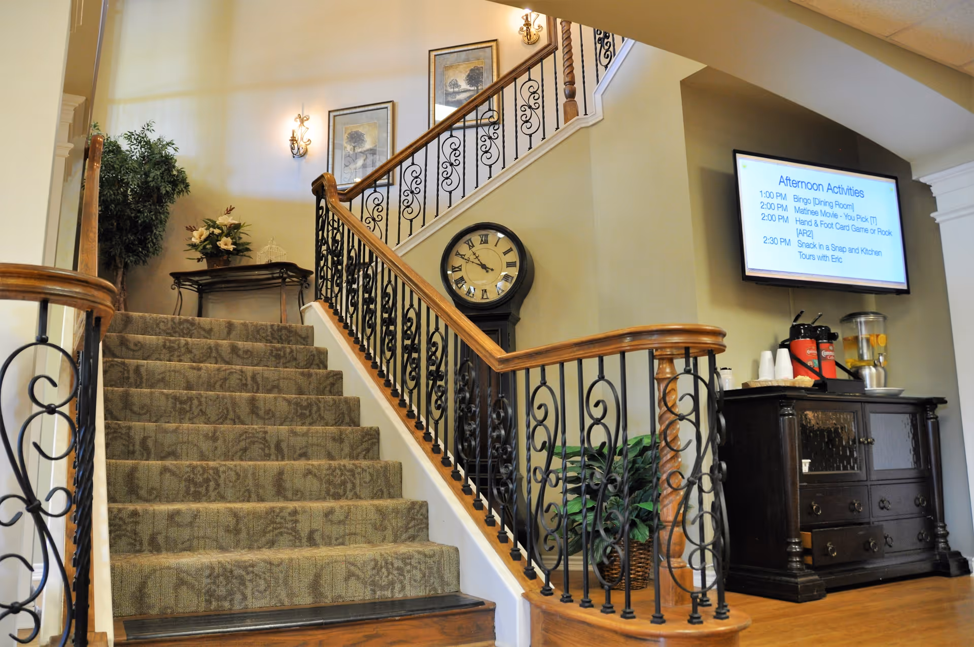 Carpeted staircase with decorative iron railing in a senior living facility lobby, featuring a large wall clock, activity screen, and a beverage station.