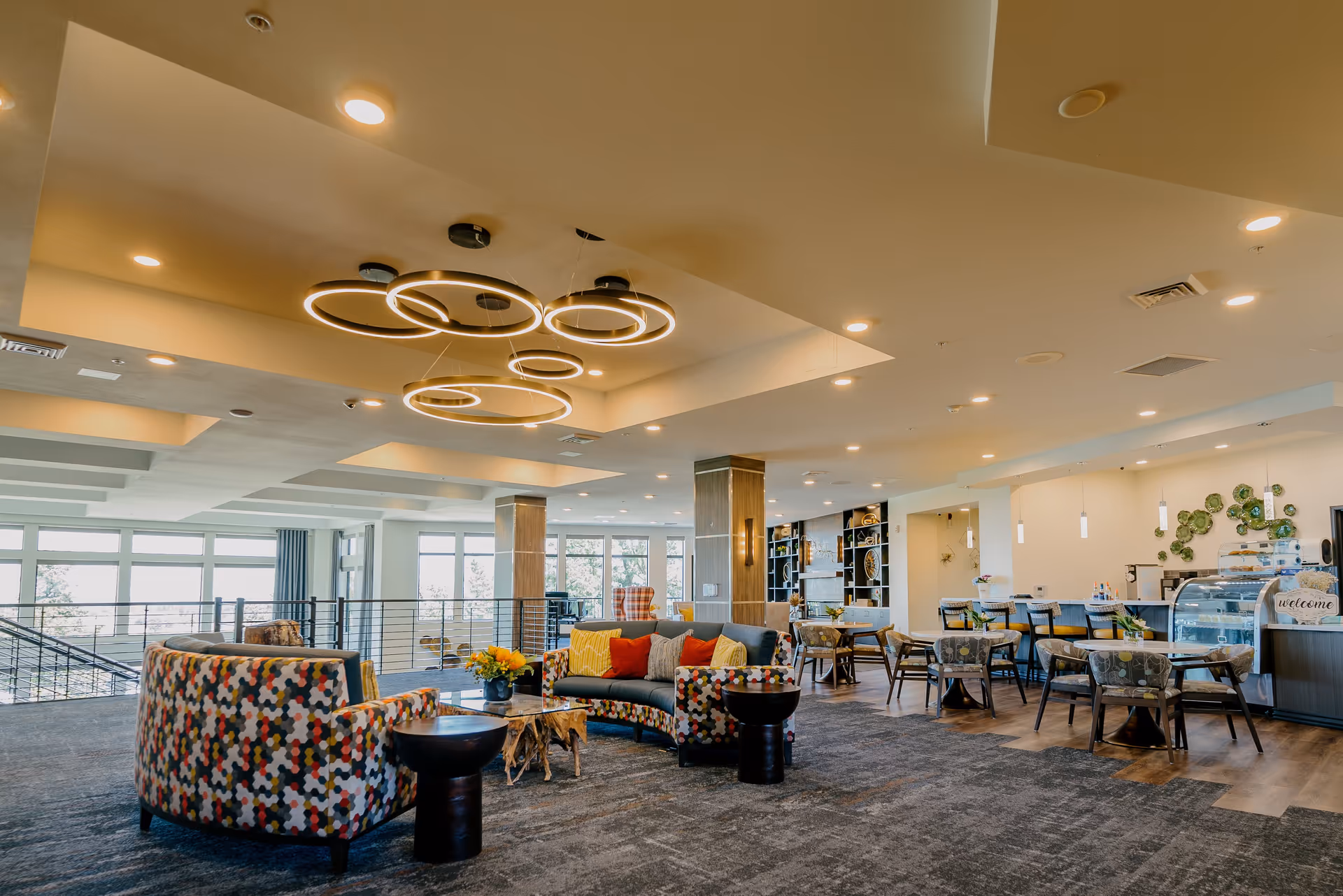 A spacious and modern common area in a senior living facility featuring colorful patterned sofas with cushions, a wooden coffee table with a flower arrangement, multiple dining tables and chairs, a display case, and contemporary circular ceiling lights. Large windows allow natural light to fill the room.