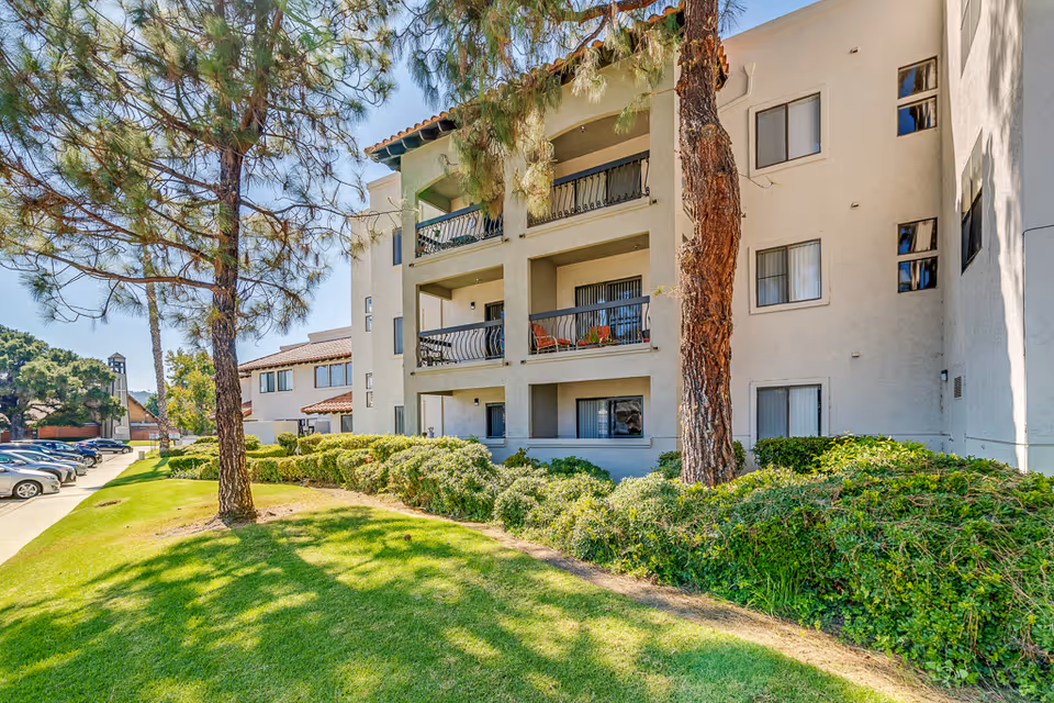 Exterior view of a multi-story senior living facility building with balconies, surrounded by green grass, bushes, and tall trees. Several parked cars are visible along the driveway on the left side under a clear blue sky.