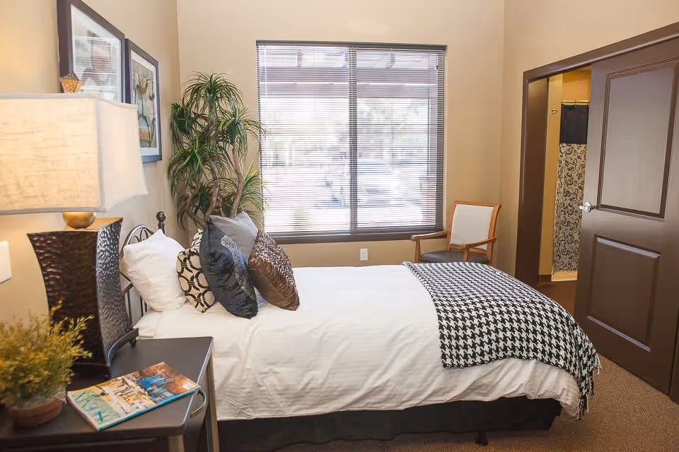 A cozy bedroom with a neatly made bed featuring multiple decorative pillows and a black and white houndstooth throw blanket. There is a bedside table with a lamp and a magazine, a potted plant, a framed picture on the wall, a large window with blinds, and a chair near the door. The door is open, revealing a glimpse of a bathroom with a patterned shower curtain.