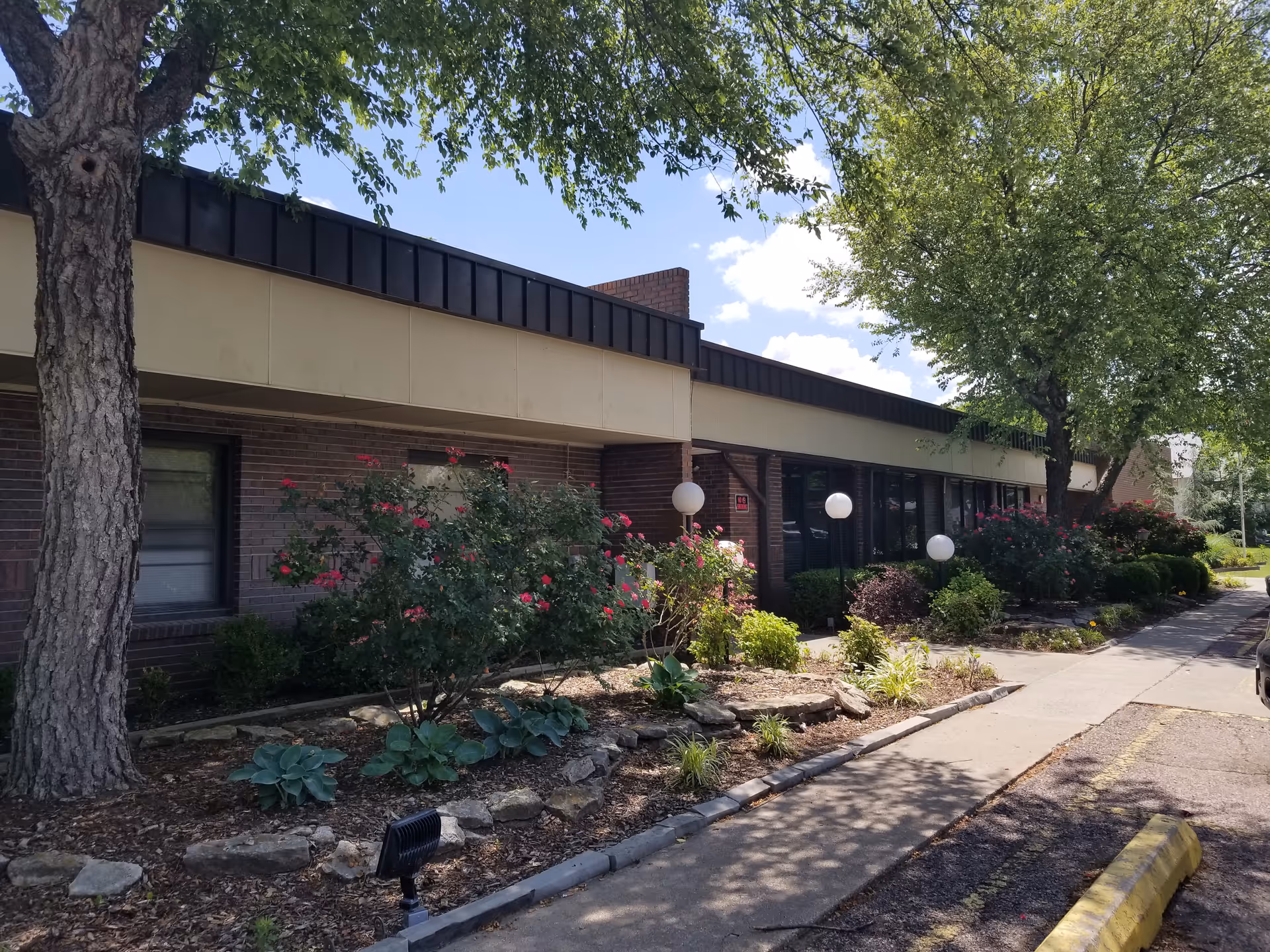 Exterior view of Ballard Nursing Center showing a single-story brick building with large windows, surrounded by landscaped garden beds with bushes, flowers, and trees under a partly cloudy sky.