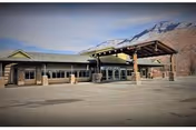 Exterior view of a single-story building with a covered entrance and large windows, set against a backdrop of mountains under a partly cloudy sky. The parking lot in front is empty.