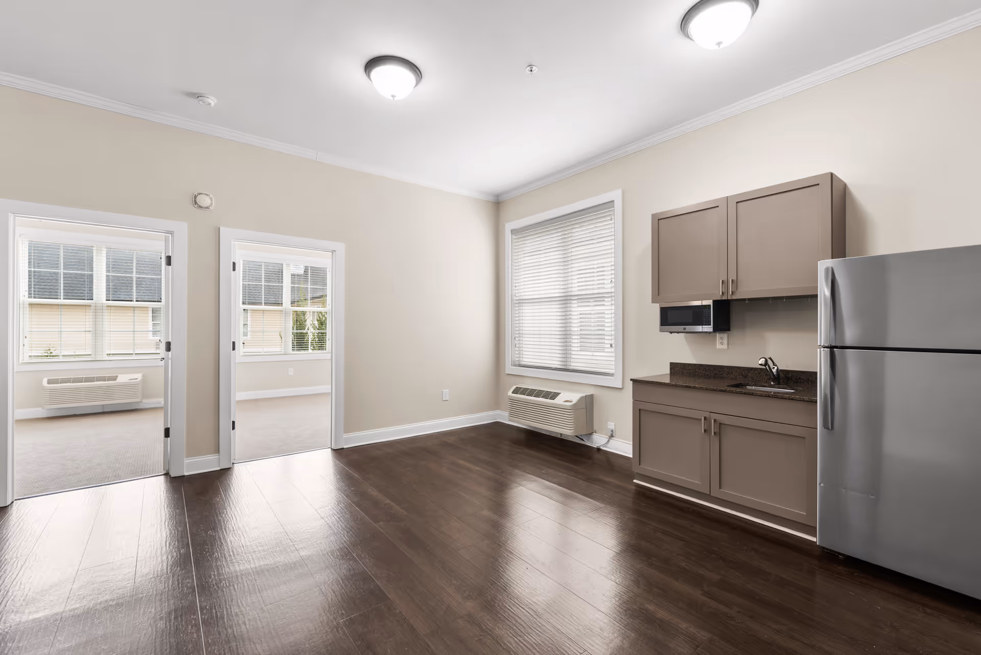 An empty interior room with dark wood flooring, beige walls, and white trim. The room features a small kitchenette with brown cabinets, a microwave, a sink, and a stainless steel refrigerator. There are two doorways leading to adjacent rooms with carpeted floors and large windows with blinds. The room is well-lit with ceiling lights and natural light from the windows.