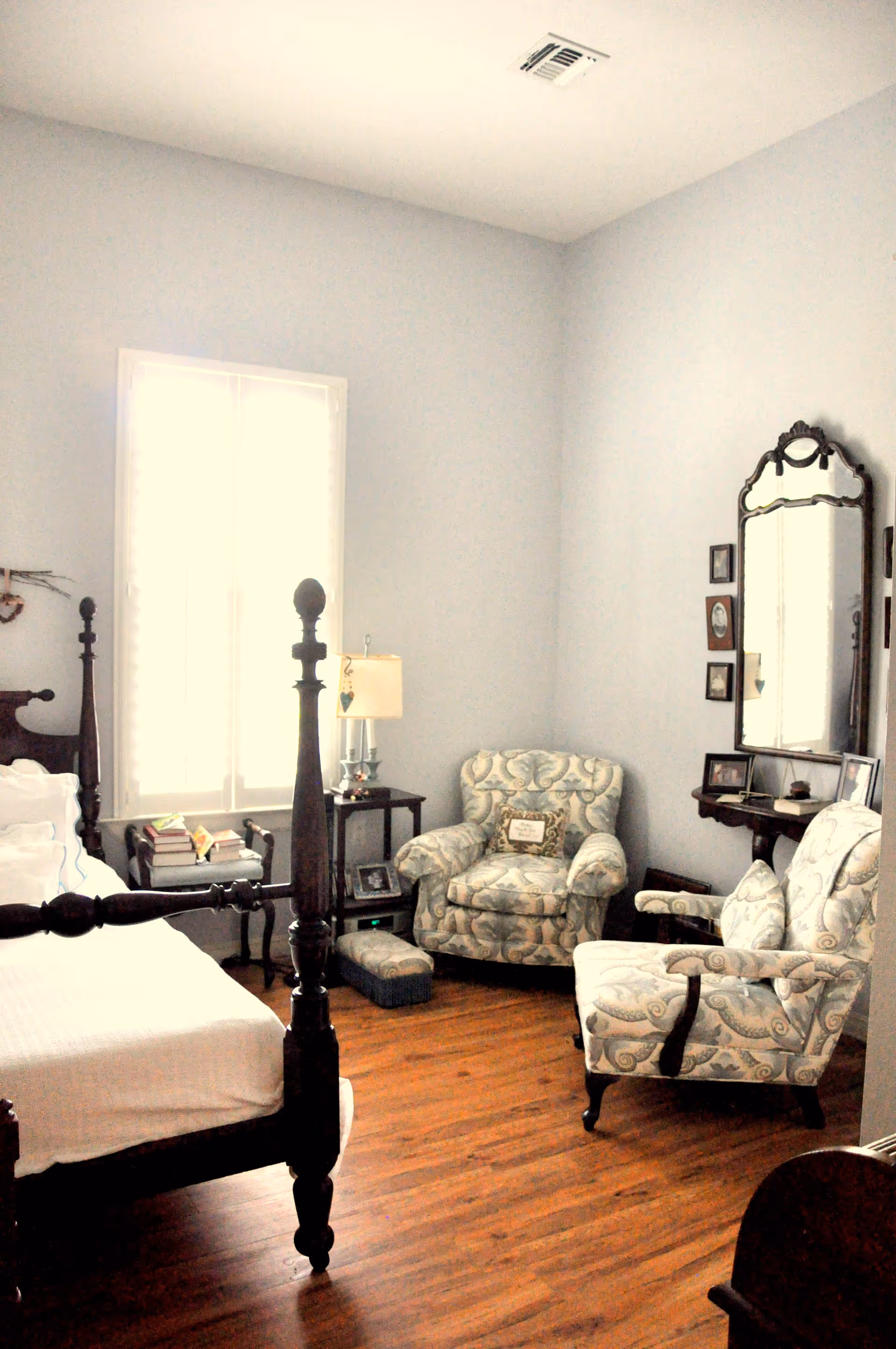 A cozy bedroom with a wooden four-poster bed on the left, a window with white shutters letting in natural light, two upholstered armchairs with a floral pattern, a small side table with a lamp, and a wall-mounted mirror with framed pictures around it. The floor is wooden and the walls are painted light gray.