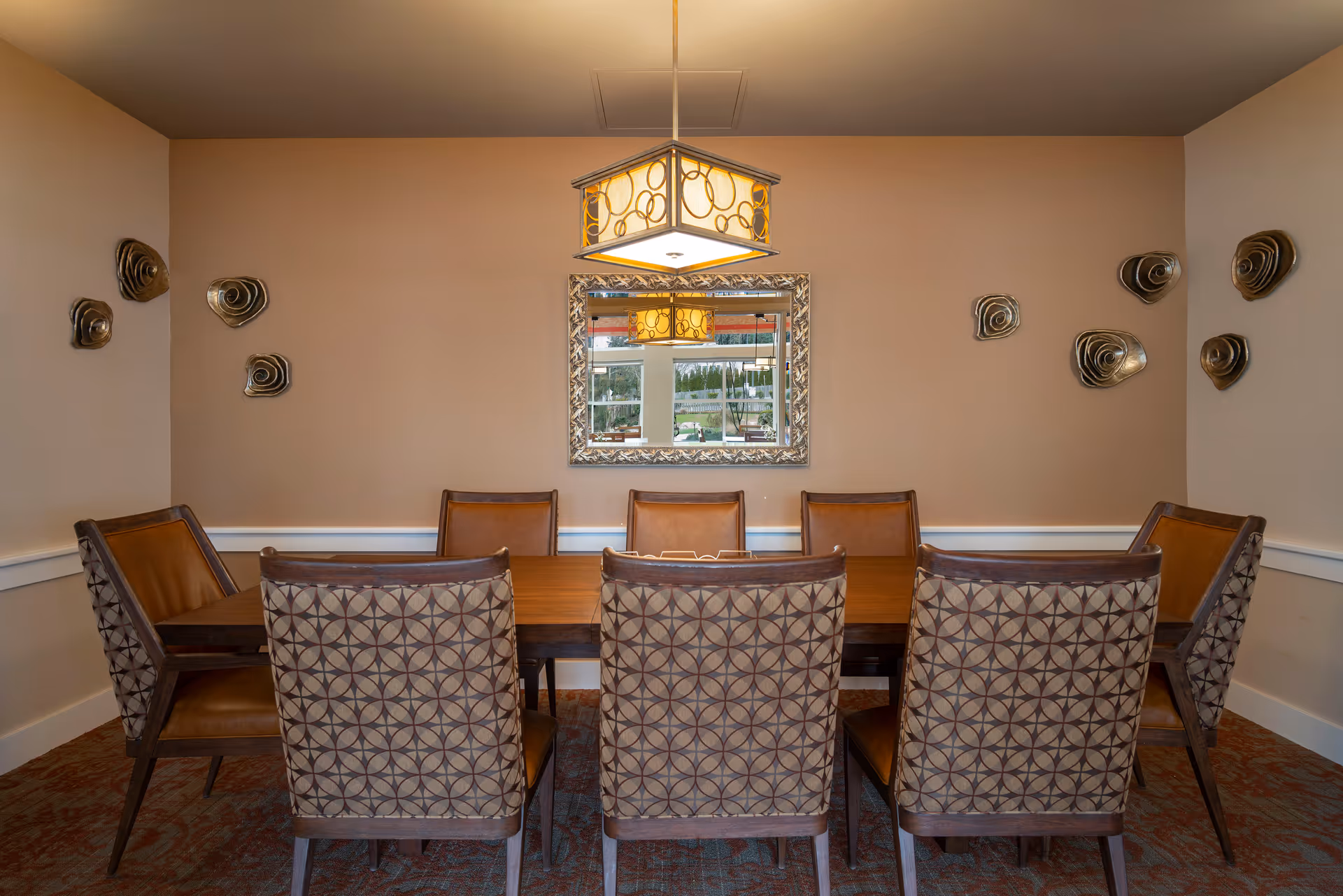 A formal dining room with a wooden table surrounded by eight patterned chairs, a decorative hanging light, wall sculptures and a mirror.