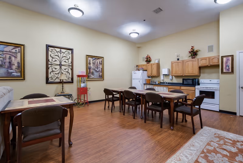 Communal dining room with several tables and chairs, a kitchenette with stove and refrigerator, wall art, and hardwood floors.