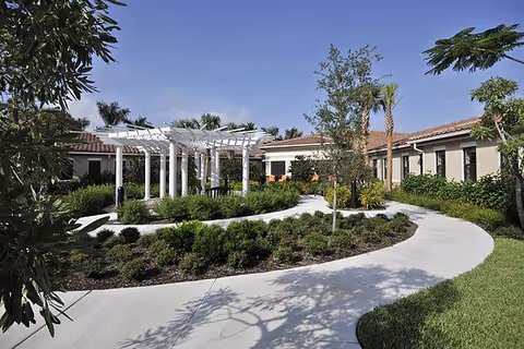 Outdoor courtyard area at Allegro Senior Living with a curved concrete walkway, landscaped garden beds, small trees, and a white pergola structure surrounded by a single-story building under a clear blue sky.