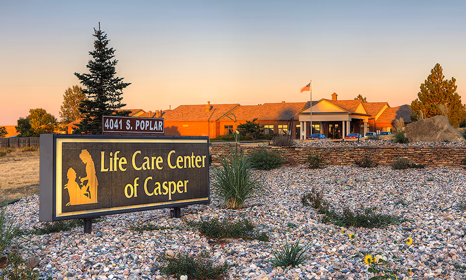 Exterior view of the Life Care Center of Casper building during sunset with a landscaped rock garden in the foreground and a sign displaying the facility's name and address, 4041 S. Poplar.
