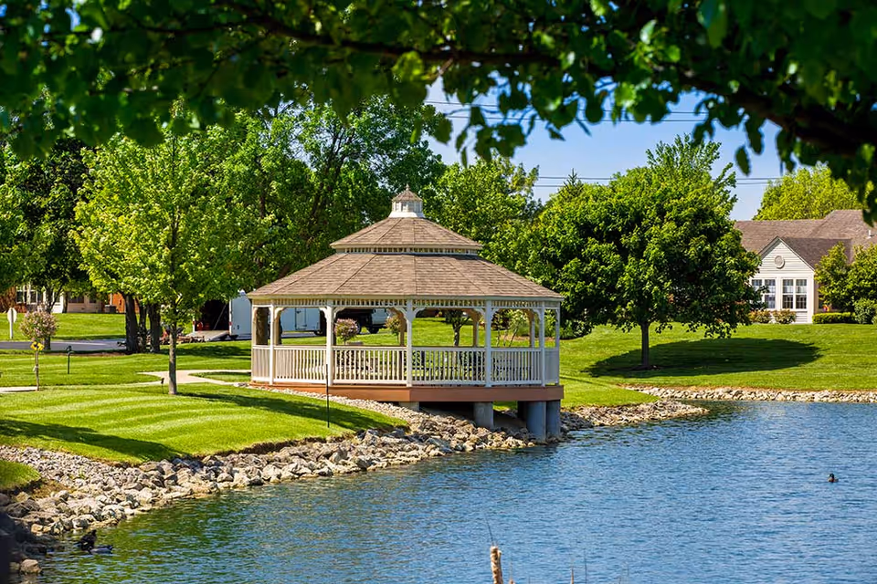 A white wooden gazebo with a shingled roof situated on the edge of a pond surrounded by green grass, trees, and a few buildings in the background under a clear blue sky.