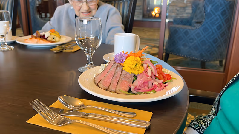 A senior woman sitting at a dining table with a plate of food in front of her, including slices of steak, pickled onions, cherry tomatoes, and edible flowers. There is a glass of water, a white mug, and silverware on a yellow napkin on the table. The background shows a cozy interior with a fireplace and upholstered chairs.