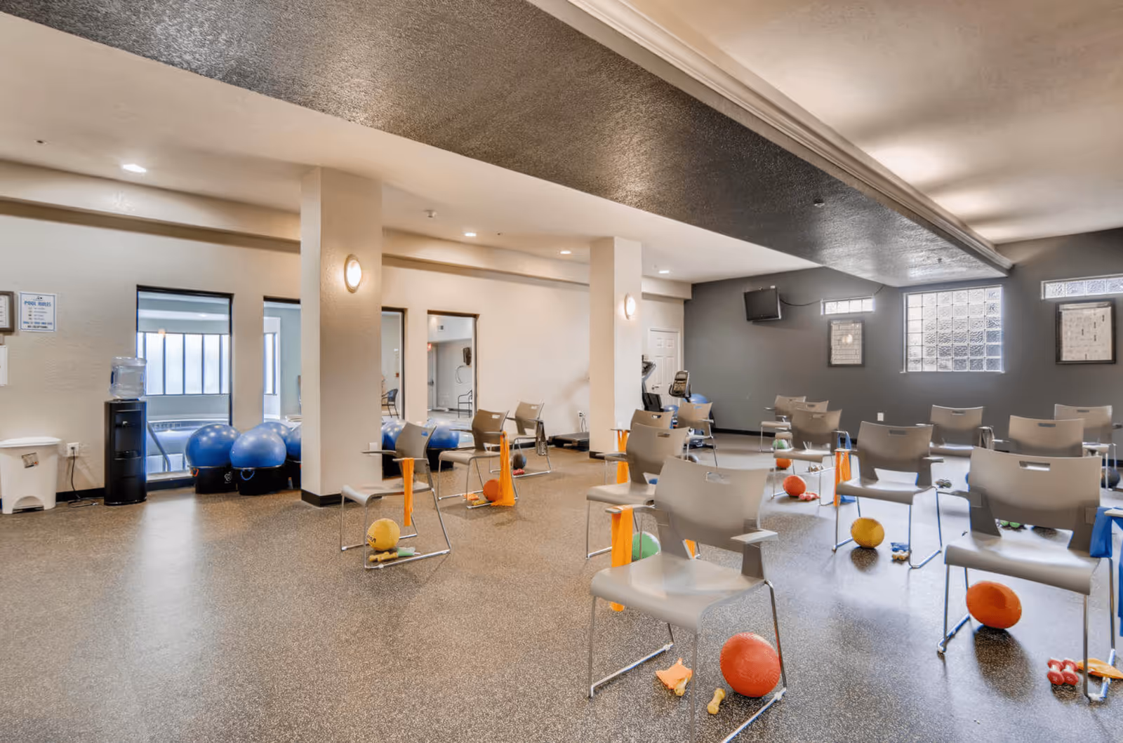 A spacious exercise room with multiple gray chairs arranged in rows, each accompanied by small exercise equipment such as balls, dumbbells, and resistance bands. The room has a textured ceiling, light-colored walls, and windows letting in natural light. There is a water cooler and exercise balls in the corner, and a wall-mounted TV on the far wall.
