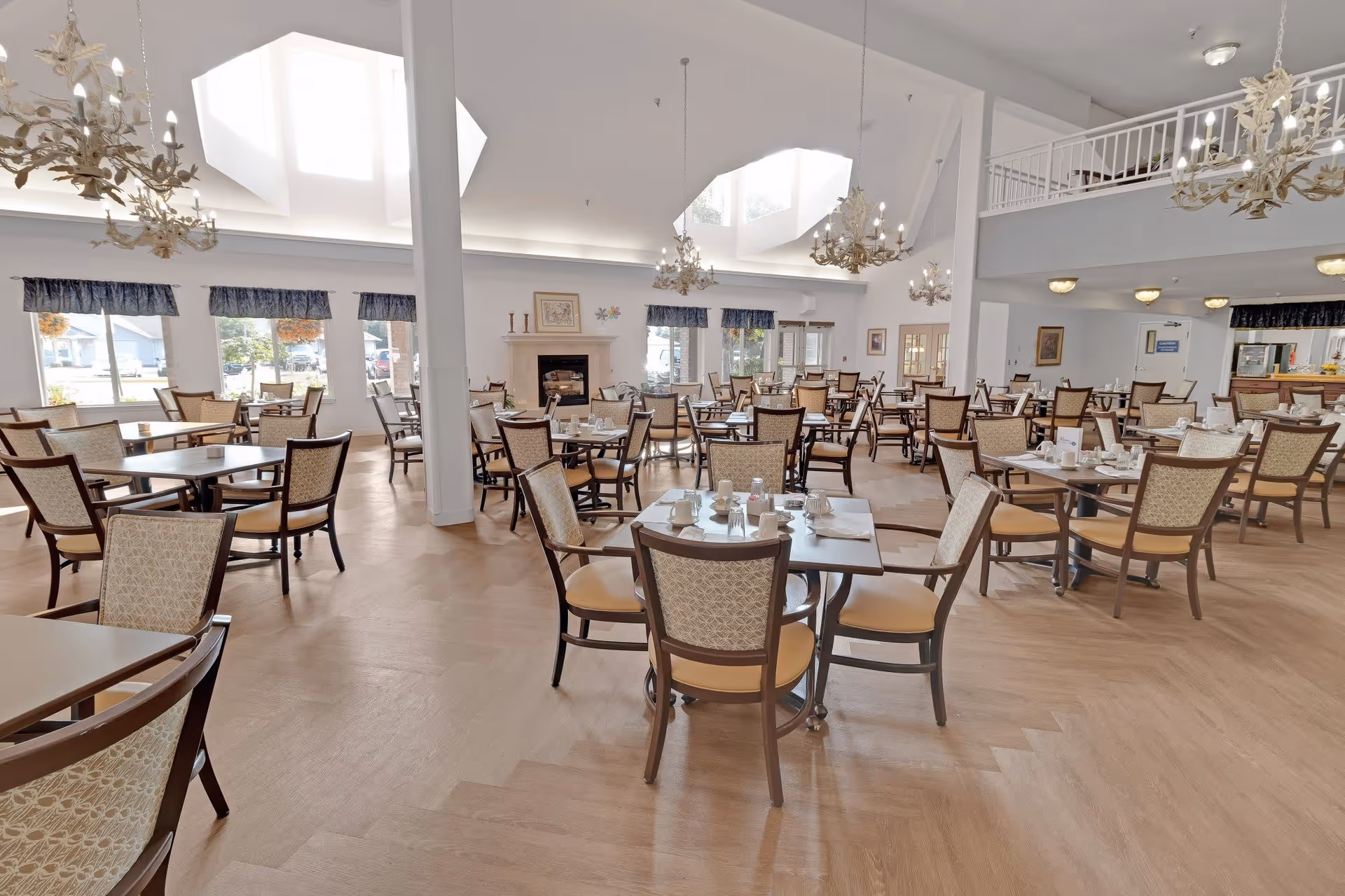 A spacious dining room with multiple tables and chairs arranged neatly. The room features large windows with blue valances, chandeliers hanging from a high ceiling with skylights, and a fireplace in the background. Tables are set with cups, saucers, and condiments, creating a welcoming atmosphere.