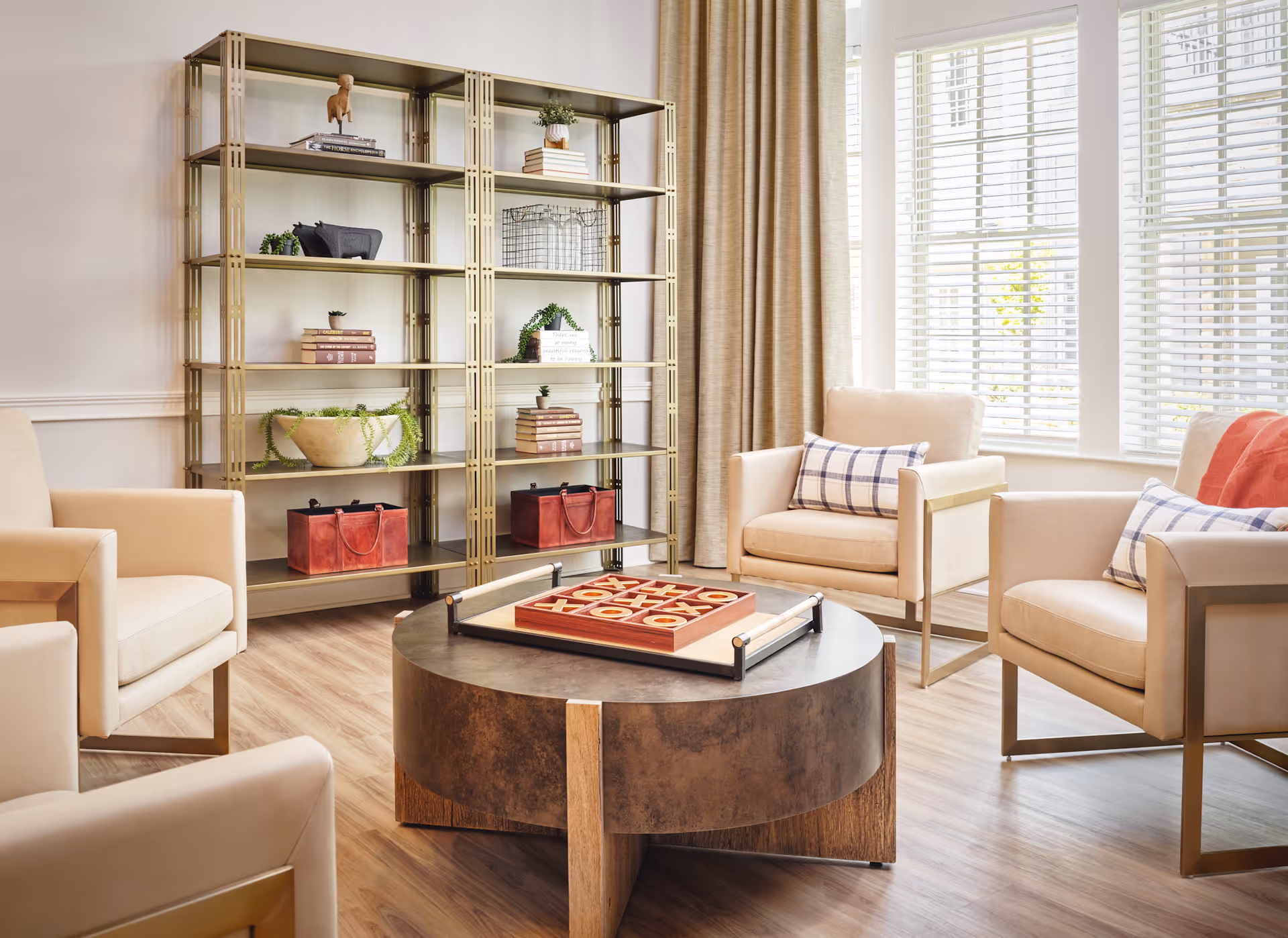 A bright and cozy living room with four beige armchairs arranged around a round wooden coffee table. On the table is a tic-tac-toe game board. Behind the chairs is a tall metal shelving unit with decorative items including books, plants, and baskets. Large windows with white blinds and beige curtains allow natural light to fill the room.