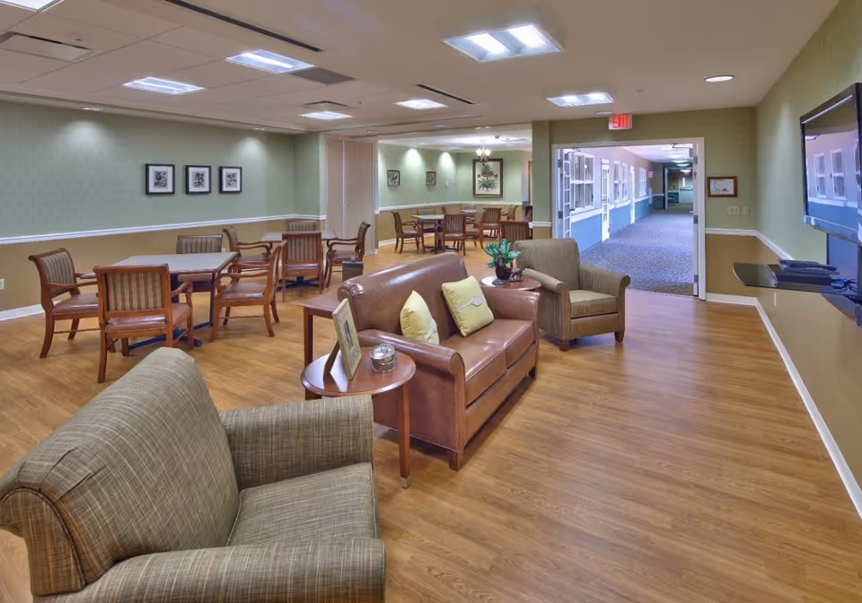 A spacious senior living common area with wooden flooring, green and beige walls, and ceiling lights. The room is furnished with a brown leather sofa with two yellow pillows, two armchairs, several wooden tables with chairs, and a flat-screen TV mounted on the wall. A hallway with blue carpet and windows is visible through an open doorway.