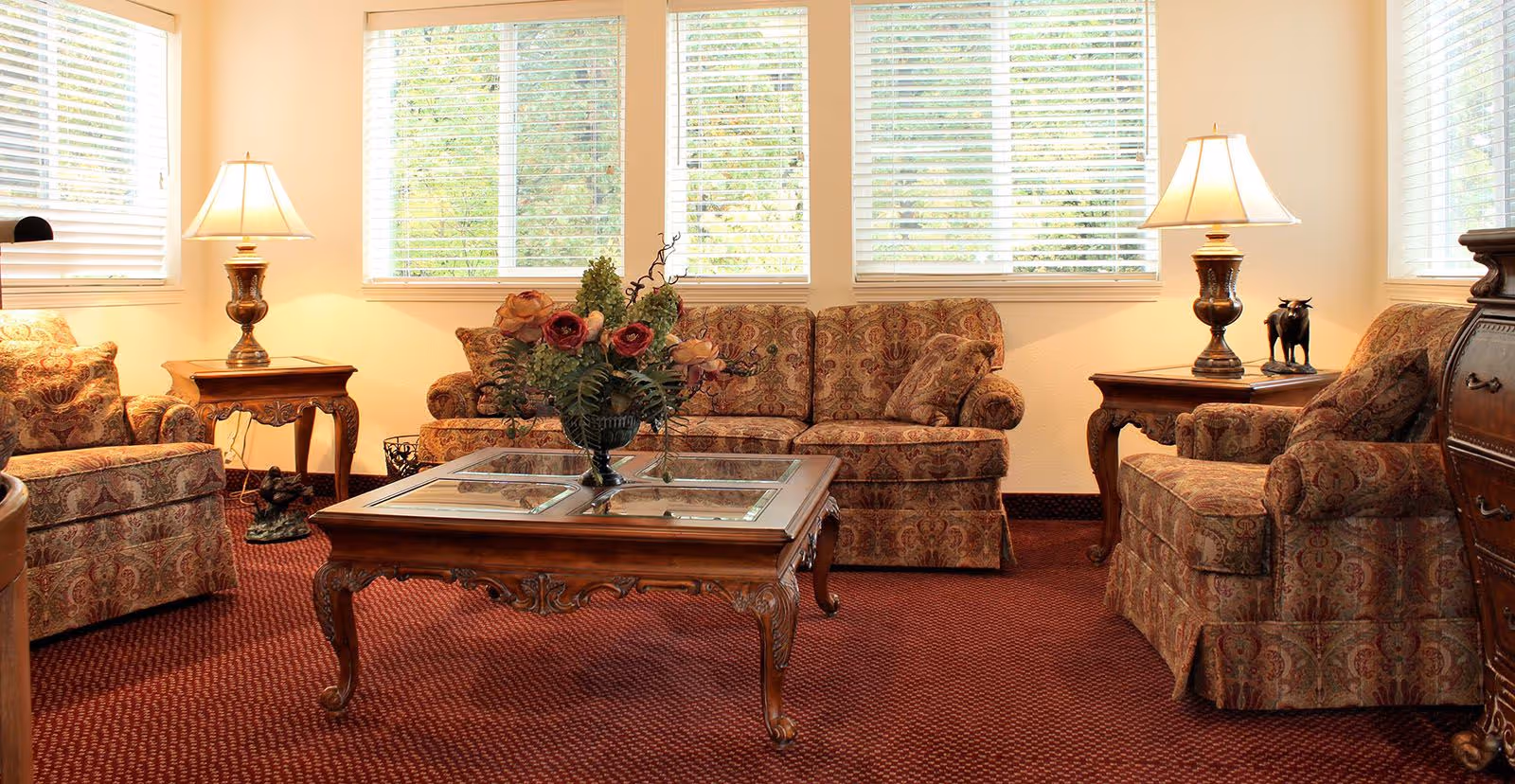 A cozy living room with patterned upholstered sofa and armchairs, a wooden coffee table with a floral arrangement, two side tables each with a lamp, and large windows with white blinds letting in natural light.