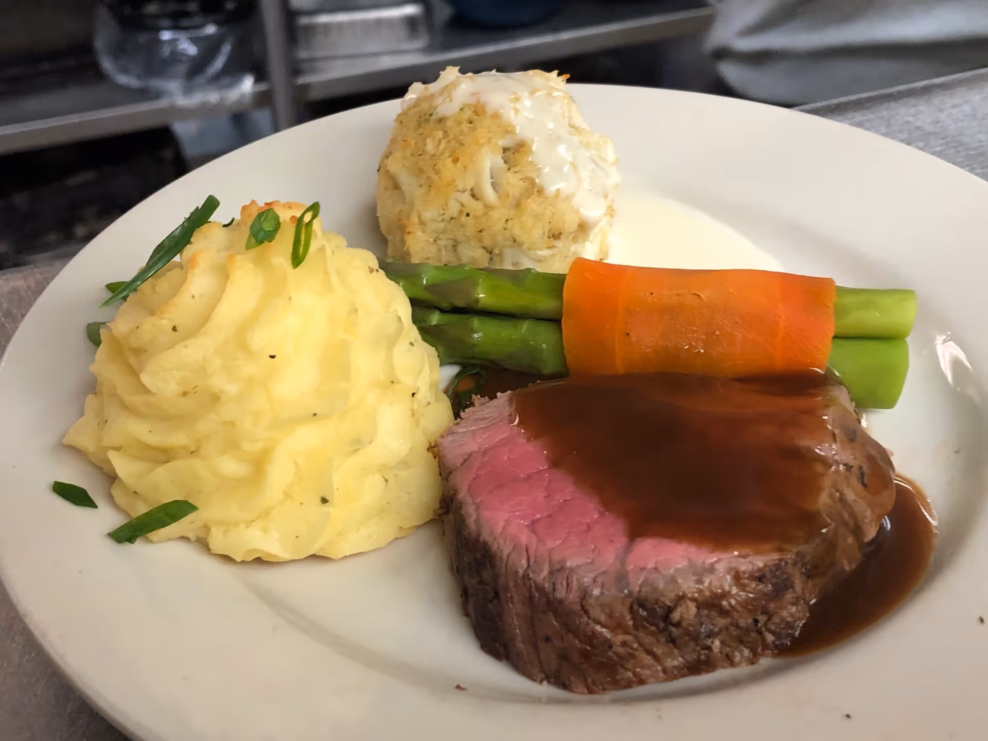 A plated meal featuring a slice of medium-rare roast beef with brown gravy, mashed potatoes garnished with chopped green onions, a crab cake with white sauce, and asparagus wrapped in a carrot strip.