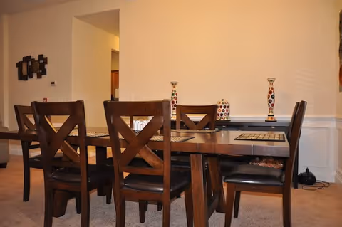 A dining room with a dark wooden table and six matching chairs with black cushions. The table has placemats and decorative items, including two tall vases with colorful patterns and a matching container. The room has beige walls and carpeted floor, with a partial view into an adjacent room.