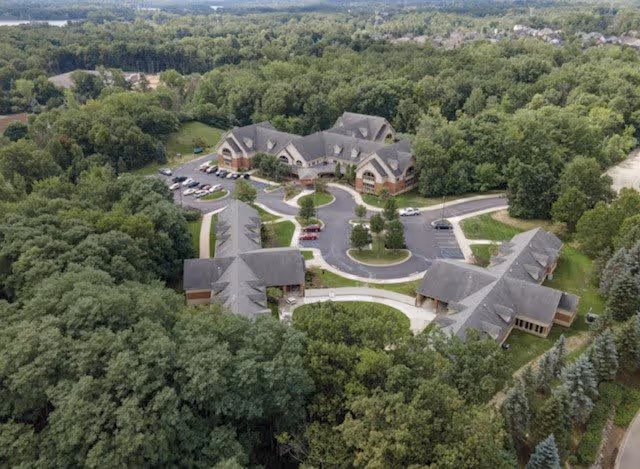 Aerial view of Addington Place of Clarkston surrounded by dense green trees. The facility consists of multiple connected buildings with gray roofs and a circular driveway with parked cars. The landscape is lush and forested, extending into the horizon.
