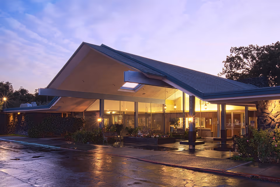 Exterior view of Garden City Healthcare Center at dusk, showing a modern building with large glass windows, a peaked roof, and warm interior lighting. The wet pavement reflects the lights, and there are plants and trees around the entrance area.