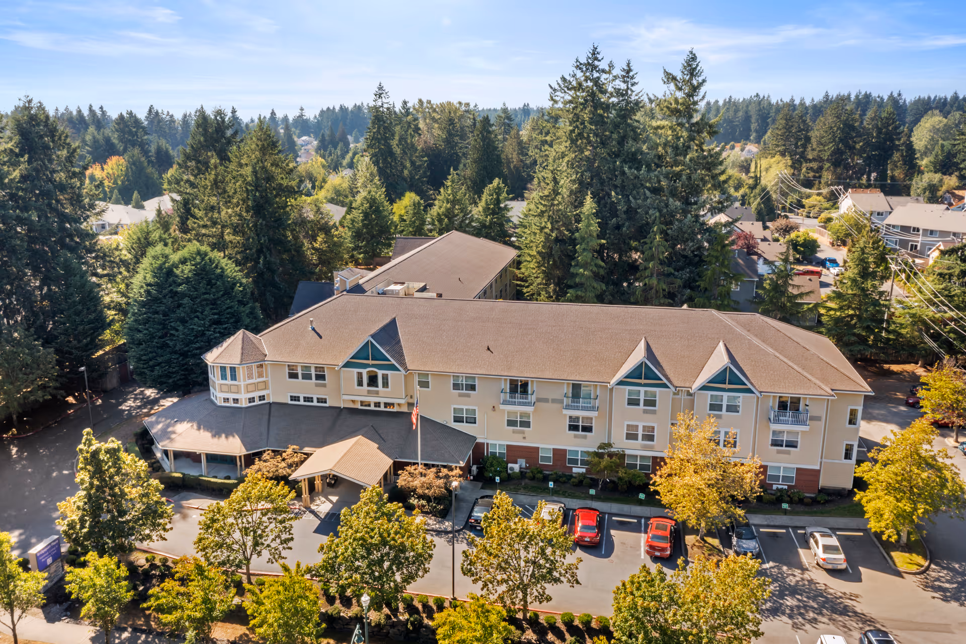 Aerial view of Bridgeport Place Assisted Living facility surrounded by trees and residential houses, showing a large multi-story building with a parking lot in front and a clear blue sky above.