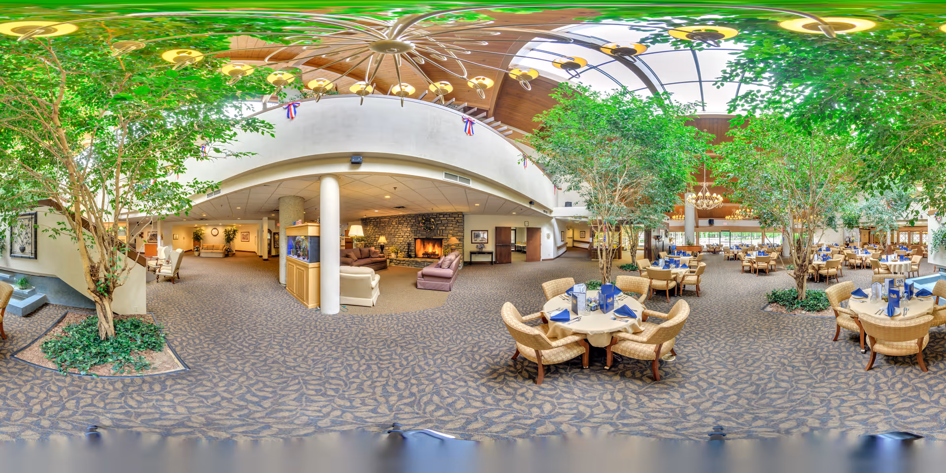 A spacious and well-lit dining area in a retirement community featuring multiple round tables set with blue napkins and menus. The room has large indoor trees, a high ceiling with skylights, and a cozy seating area with sofas and a stone fireplace. The carpet has a leaf pattern, and there are chandeliers and ceiling fans hanging from the ceiling.