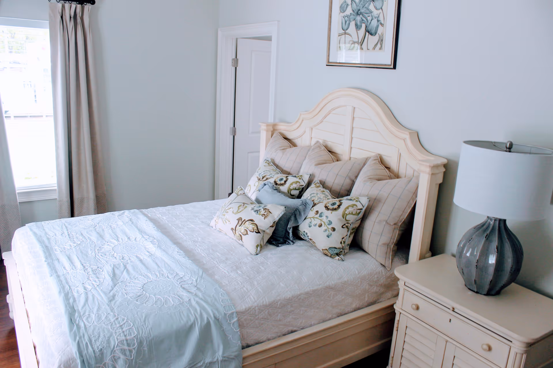 Bright bedroom featuring a cream wooden bed with patterned pillows, a nightstand with a lamp, and a window with curtains.