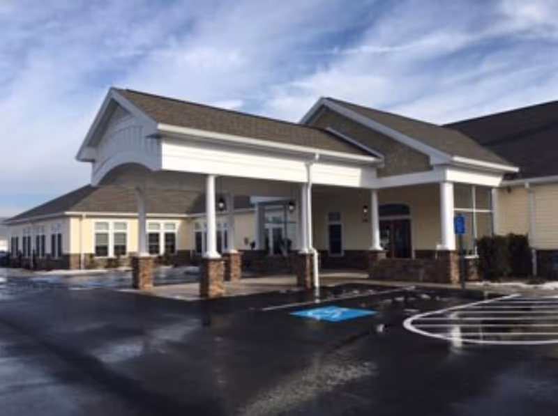 Exterior view of a single-story building with a covered entrance supported by white columns and brick bases, a parking lot with a handicap parking space in front, and a partly cloudy sky above.