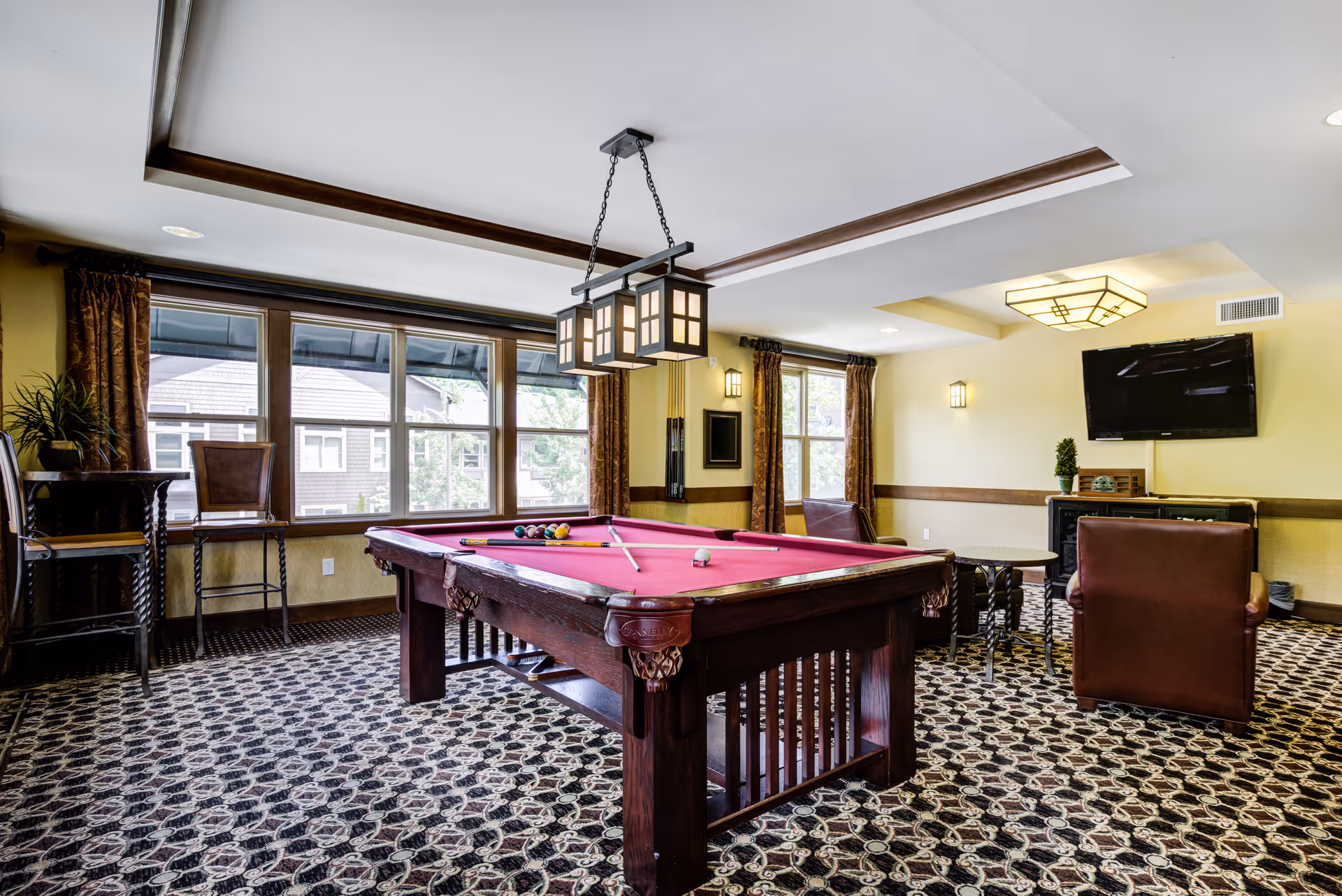 Bright communal game room with a red-felt pool table, seating, large windows, and a wall-mounted TV.