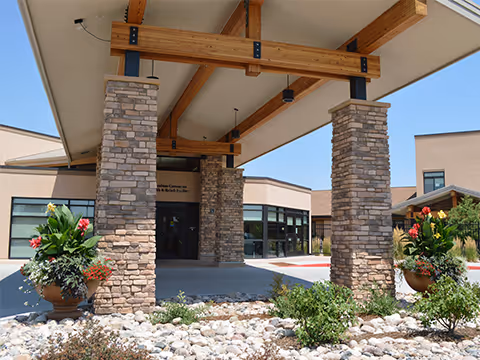 Entrance of Columbine Commons Health & Rehab Facility featuring a covered drop-off area supported by stone pillars with wooden beams. There are large planters with flowers and landscaping with rocks and shrubs in the foreground.