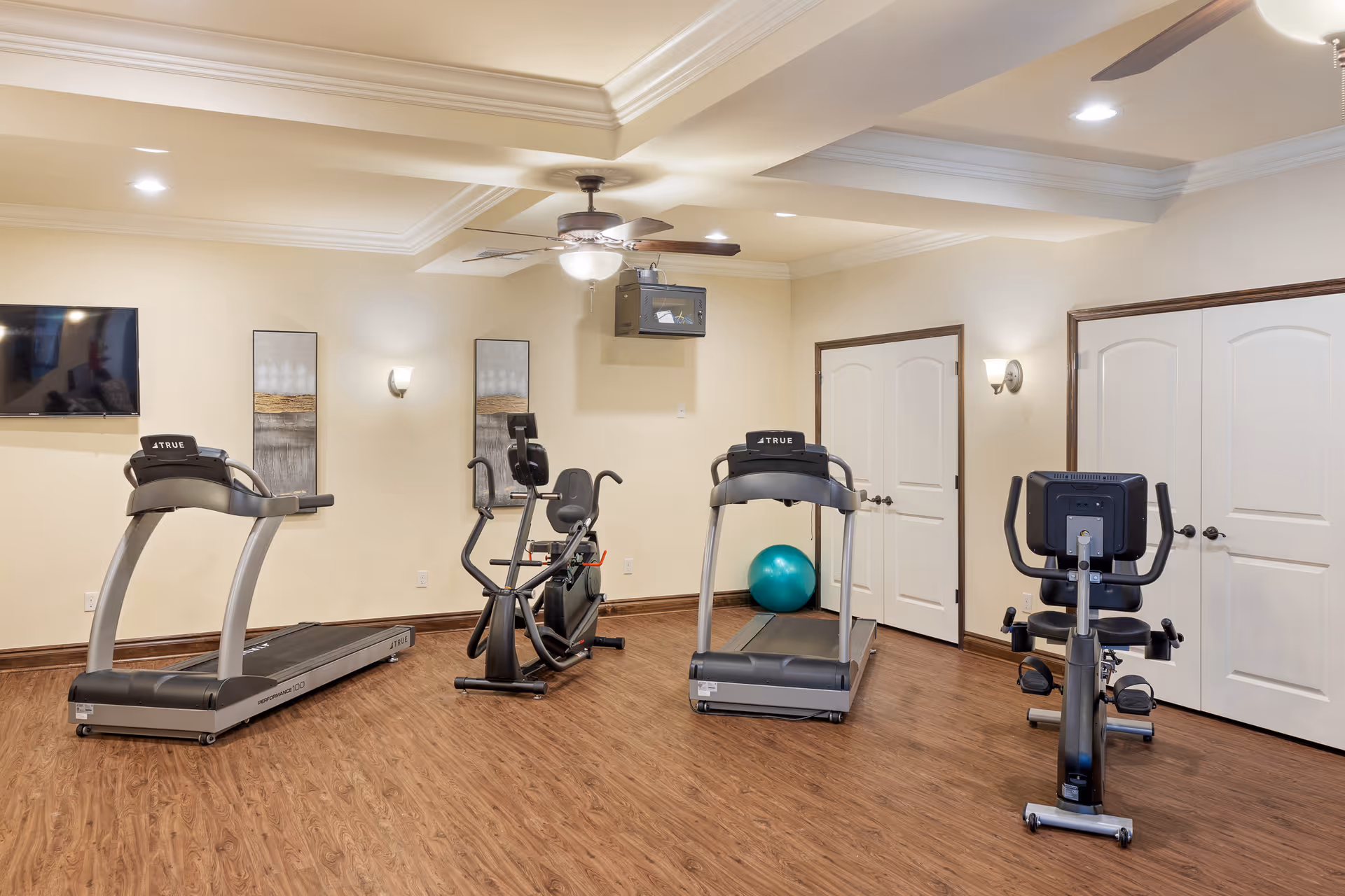 A fitness room with wooden flooring and cream-colored walls, featuring two treadmills, two stationary exercise bikes, a ceiling fan with light, a wall-mounted TV, two decorative wall panels, and a teal exercise ball in the corner.