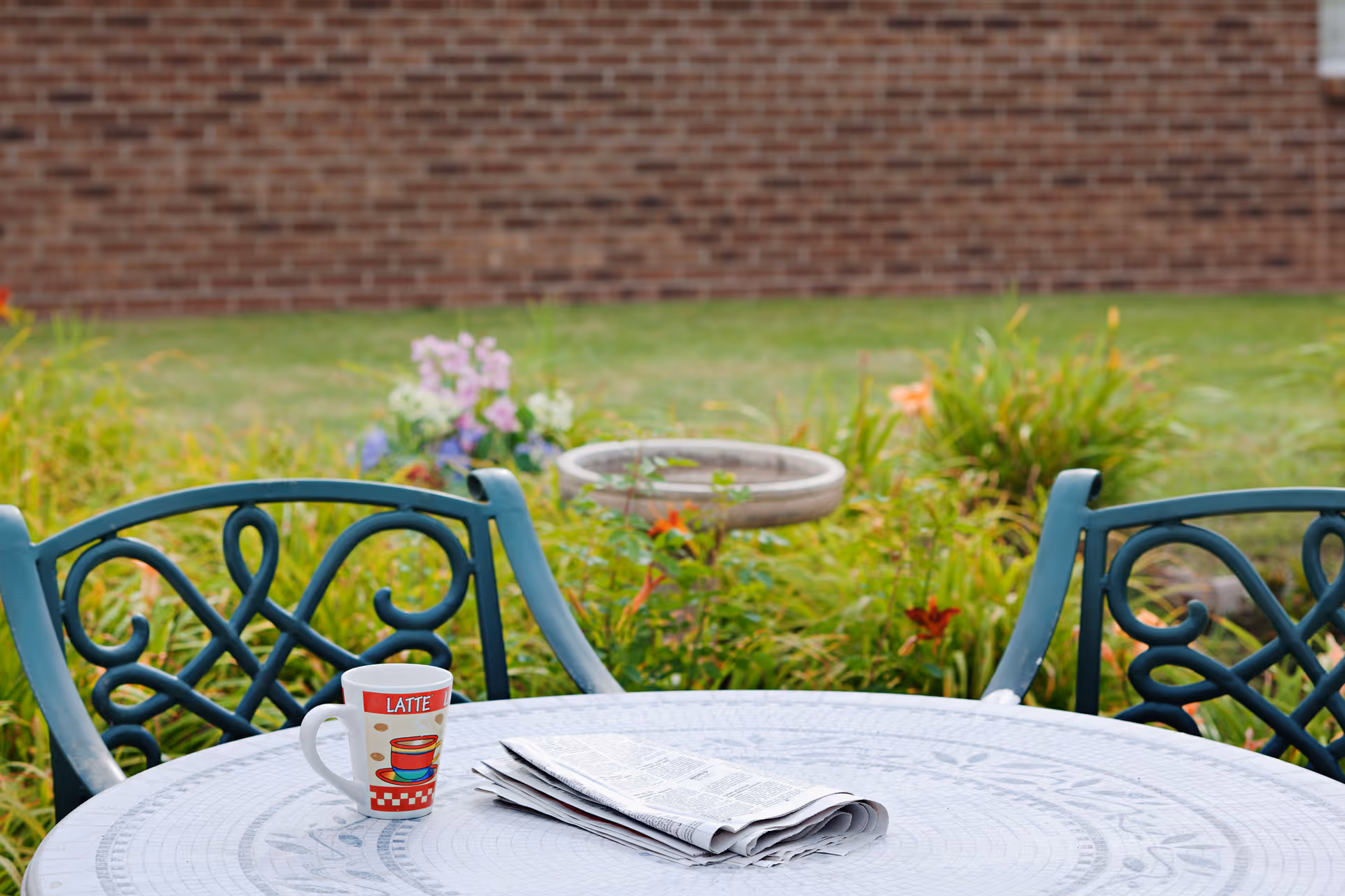 Outdoor patio area with a round metal table featuring a folded newspaper and a coffee mug labeled 'LATTE'. Two decorative metal chairs are positioned around the table. In the background, there is a garden with green plants and flowers, and a brick wall.